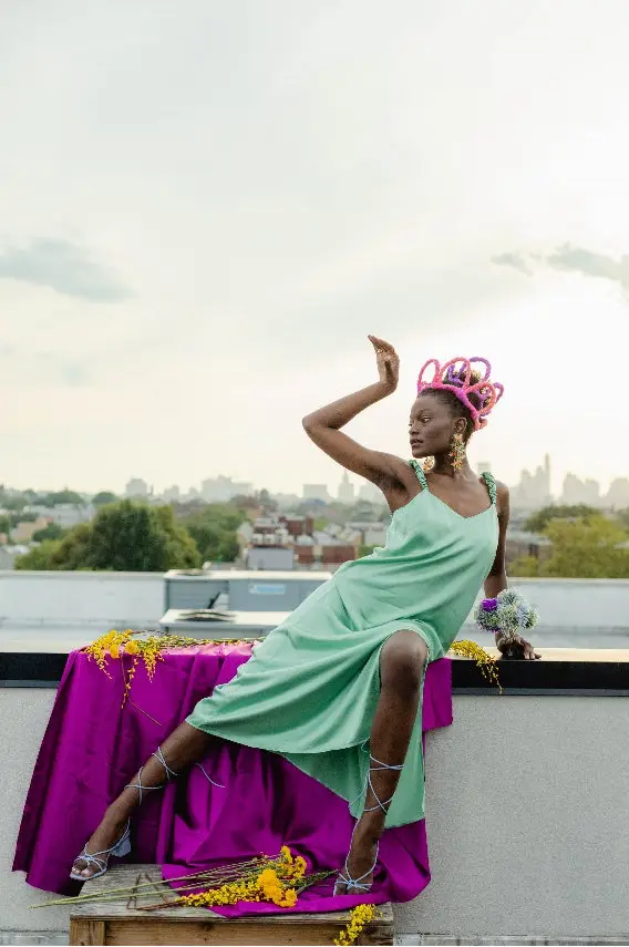 Photo of a woman wearing a green dress, pink abstract crown, posing on a purple blanket on a rooftop. 